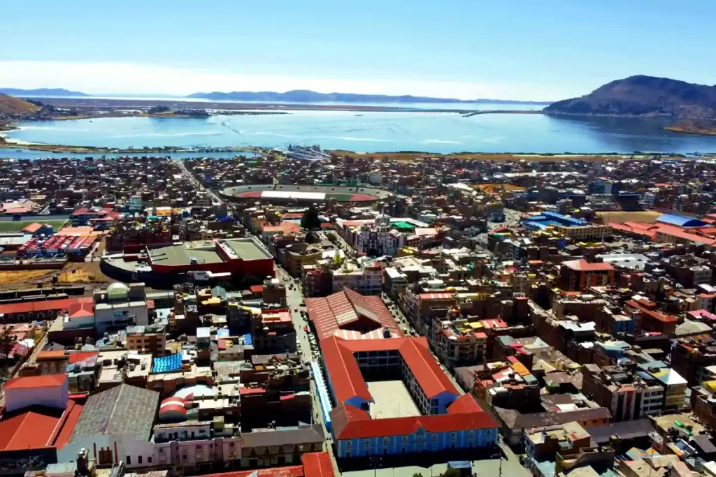 Vista panorámica de la ciudad de Puno con el Lago Titicaca al fondo, mostrando edificaciones urbanas y cerros andinos.