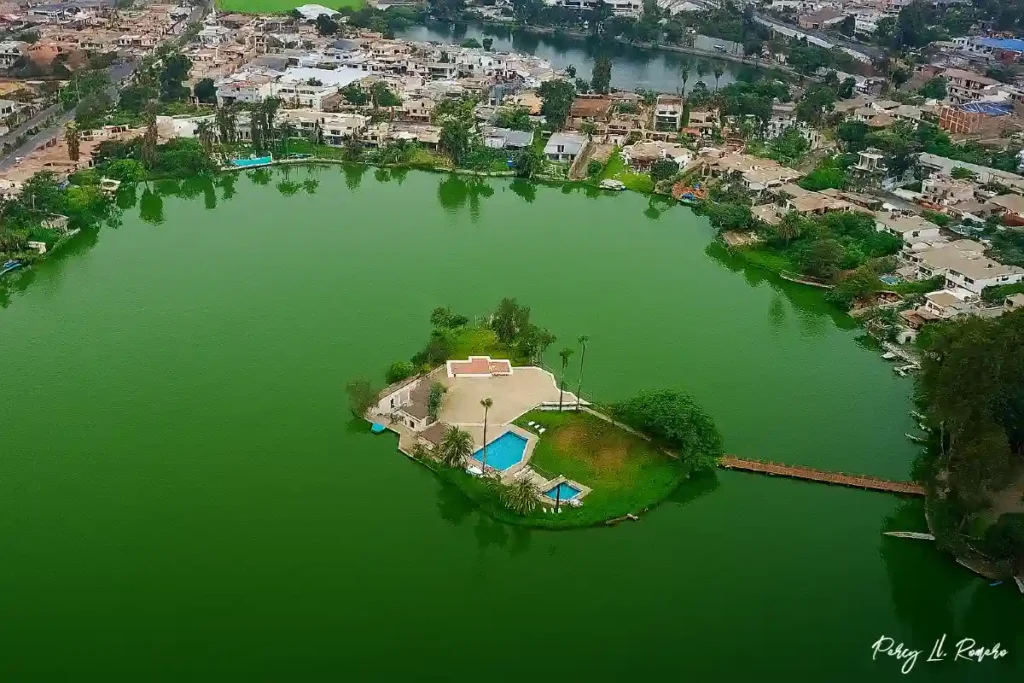 Vista aérea de la laguna La Mellicera en Chilca, Cañete, capturada en alta resolución durante el día, mostrando su isla central con piscina, rodeada de agua verde esmeralda y casas residenciales al fondo.