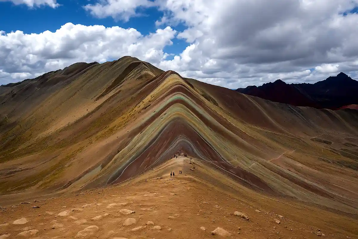 Vista panorámica de la Montaña de 7 Colores, también conocida como Vinicunca, en Cusco, con turistas ascendiendo su sendero bajo un cielo parcialmente nublado.