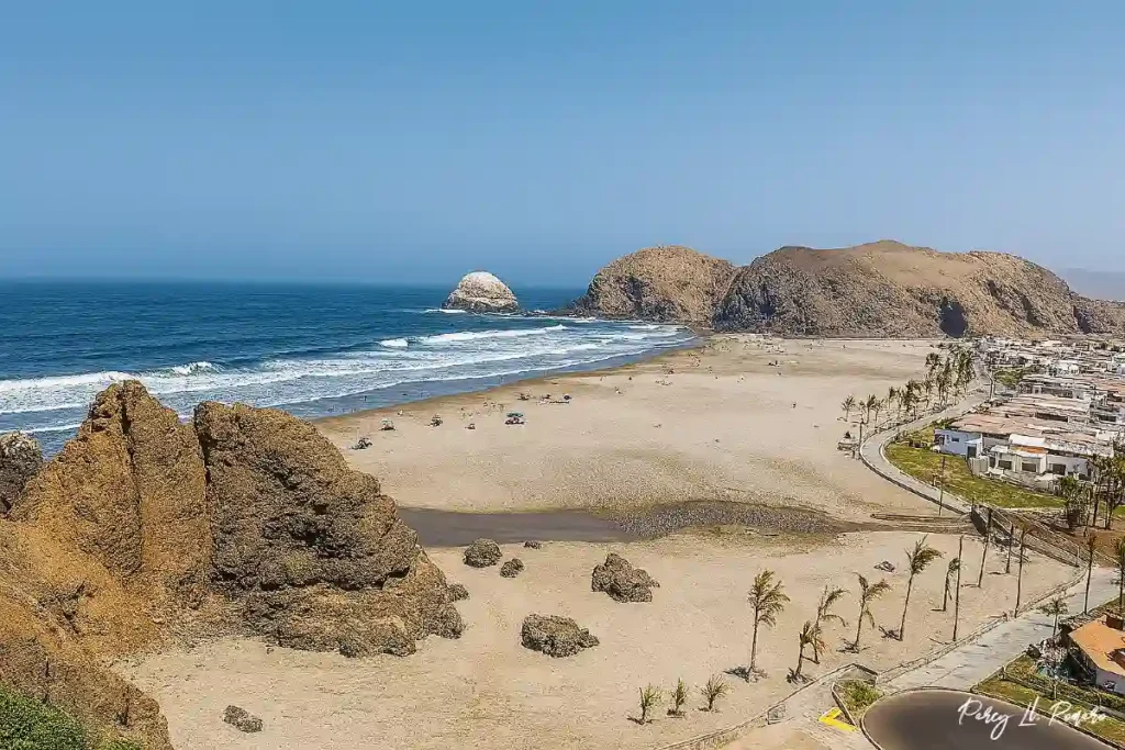 Vista panorámica de la playa Las Totoritas en Mala, Cañete, que muestra una extensa franja de arena clara, formaciones rocosas a la izquierda, mar azul con pequeñas olas y un conjunto de casas junto a una vía asfaltada en el lado derecho, todo bajo un cielo despejado y soleado.