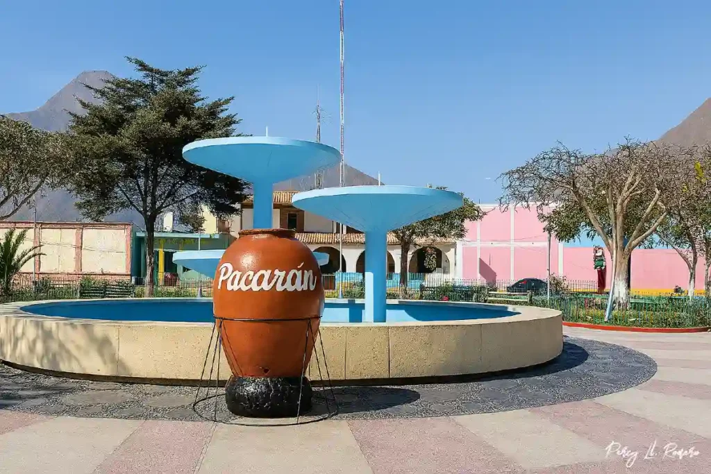 Plaza central del pueblo de Pacarán en Cañete, con una gran tinaja de barro con el nombre "Pacarán" al frente de una fuente celeste, rodeada de árboles, edificios coloridos y montañas al fondo, en un día soleado.