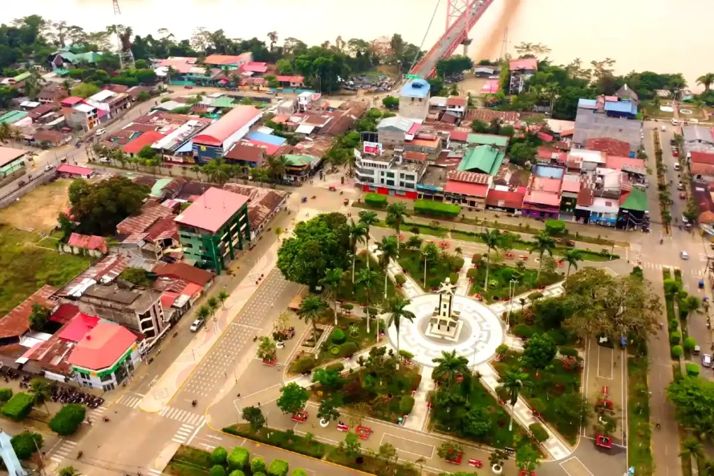 Vista aérea de Puerto Maldonado con su Plaza de Armas, rodeada de palmeras, viviendas coloridas y el puente colgante sobre el río Madre de Dios.